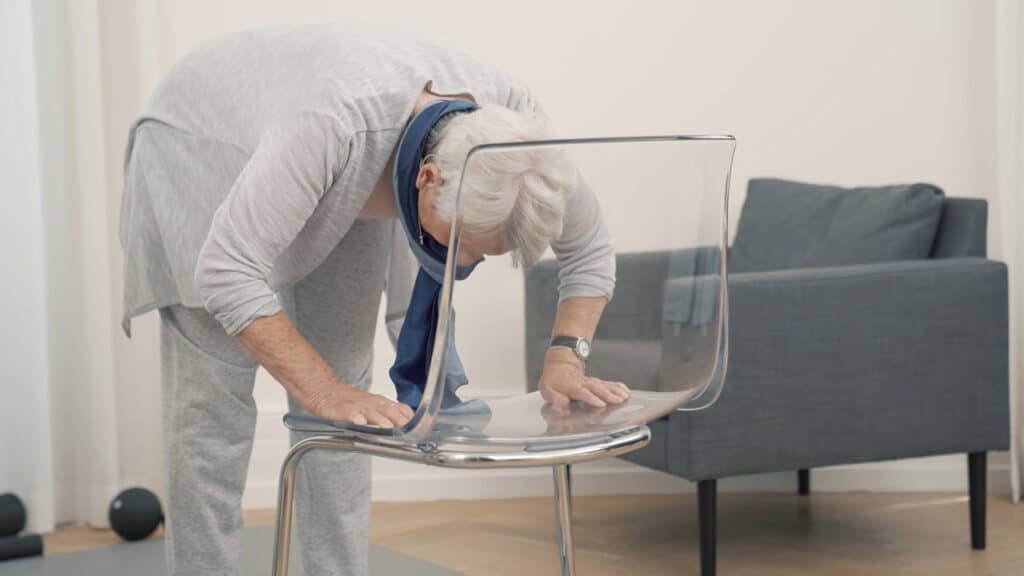 Eine ältere Dame steht vor einem Stuhl und beugt ihren Oberkörper mit geradem Rücken nach vorne.