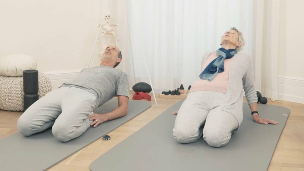 Schmerzspezialist Roland Liebscher-Bracht und seine Mutter Ruth sitzen jeweils auf ihrer grauen Übungsmatte: Sie befinden sich im Fersensitz und lehnen ihre Oberkörper nach hinten.