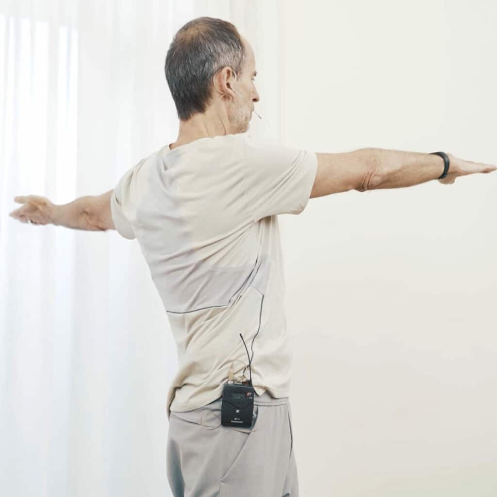 Schmerzspezialist Roland Liebscher-Bracht steht hüftbreit und er dreht seinen Oberkörper von seinen Armen geführt zur linken Seite auf.