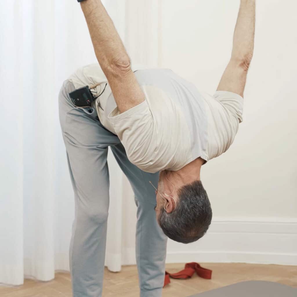 Schmerzspezialist Roland Liebscher-Bracht steht hüftbreit, mit leicht gebeugten Knien da und lässt seinen Oberkörper nach vorne und unten fallen. Beide Arme hebt er gestreckt in nach oben.