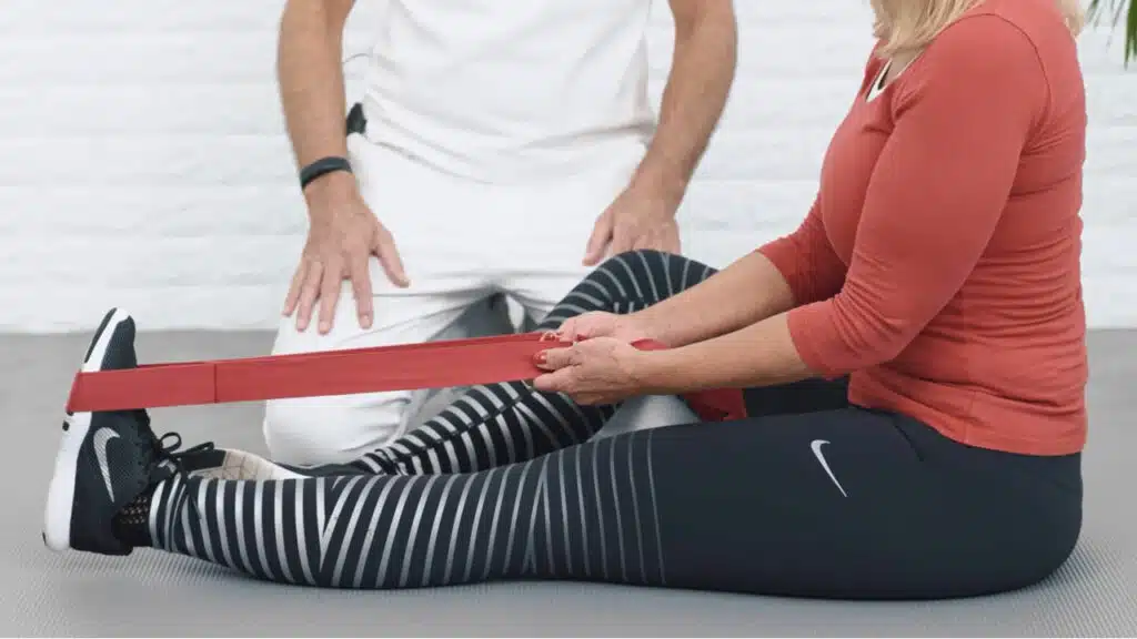 Eine Frau sitzt auf dem Boden. Ihr linkes Bein liegt lang ausgestreckt vor ihr, das rechte ist leicht aufgestellt. Die rote Übungs-Schlaufe liegt um ihren linken Vorfuß. Sie hält sie gespannt, um ihren Oberkörper mit geradem Rücken nach vorne ziehen zu können.