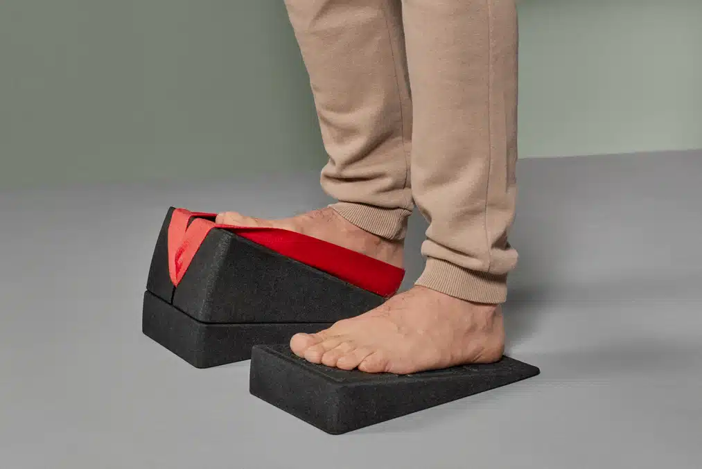 Zu sehen sind die zwei Unterschenkel eines jungen Mannes und seine zwei Füße, die jeweils auf Keilen des Liebscher & Bracht Knieretters platziert sind. Links steht er auf einem 9-Grad-Keil, rechts sind der andere 9-Grad-Keil sowie der 18-Grad-Keil aufeinandergestapelt worden. Auf diesem Höhenwinkel von 27 Grad steht sein Fuß in der roten Trittschlaufe, um nicht abzurutschen.