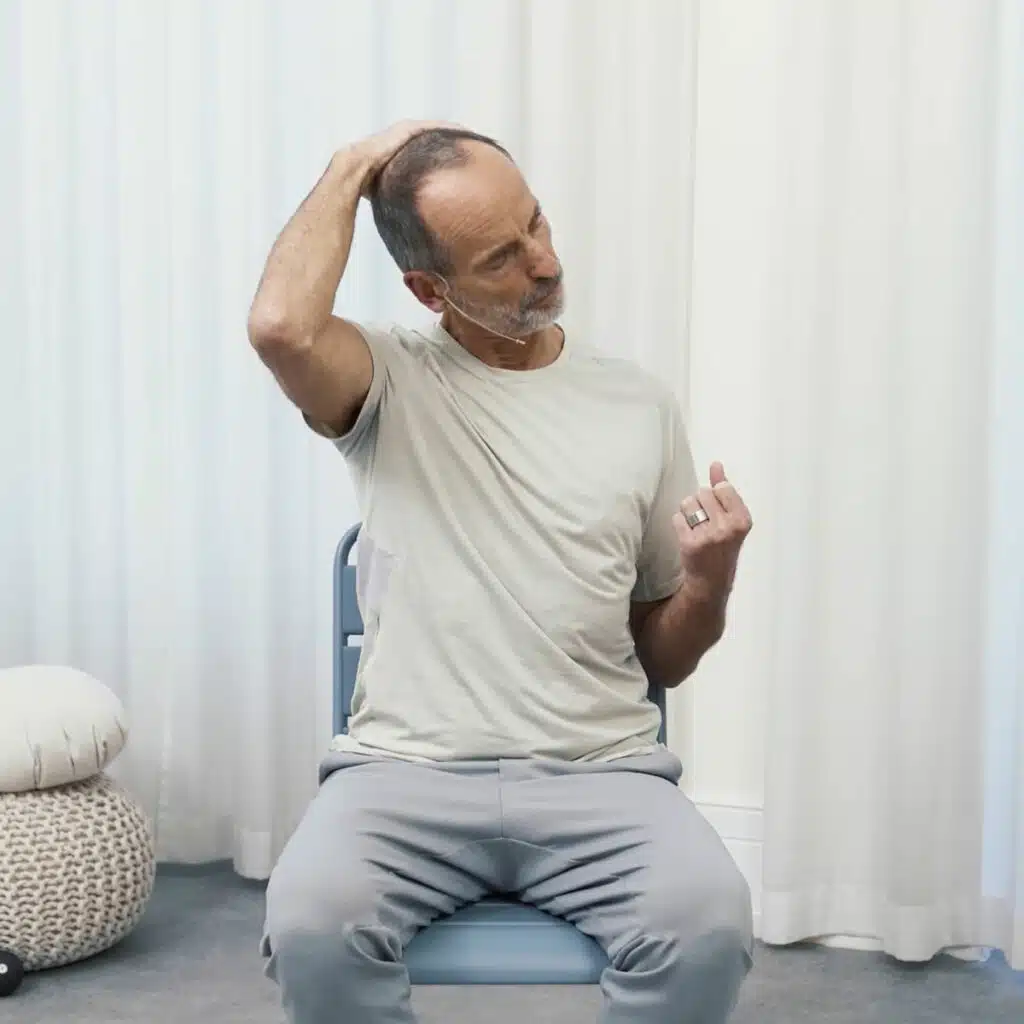 Roland Liebscher-Bracht sitzt aufrecht auf einem Stuhl. Seinen linken Arm hat er angewinkelt, die Hand zu einer Faust gebildet. Er zieht die Schulter runter, während er mit seiner rechten Hand an seinen Kopf greift und diesen nach vorne und unten zieht.