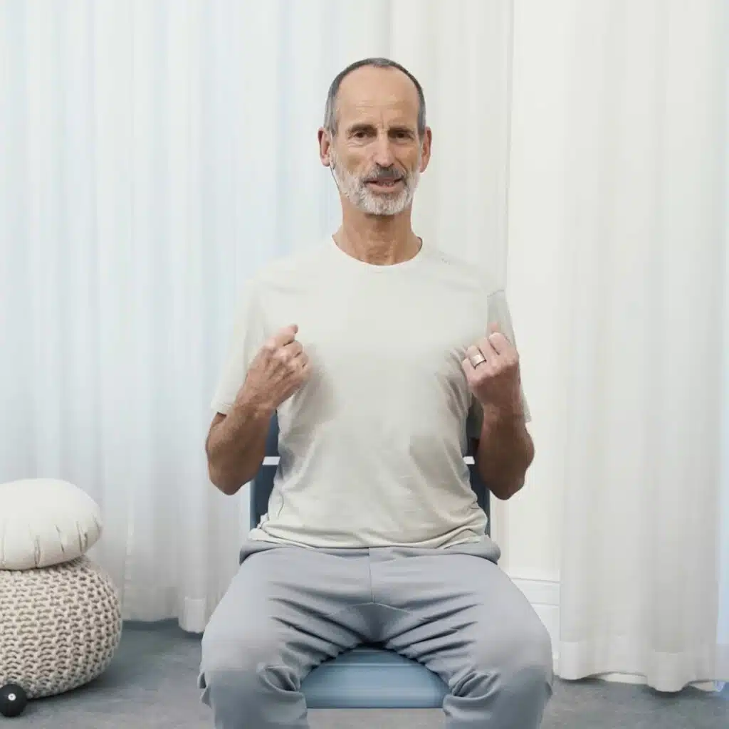 Roland Liebscher-Bracht sitzt aufrecht auf einem Stuhl. Seine Arme hat er angewinkelt. Die Hände sind leicht zu Fäusten geballt. Er zieht die Schultern runter.