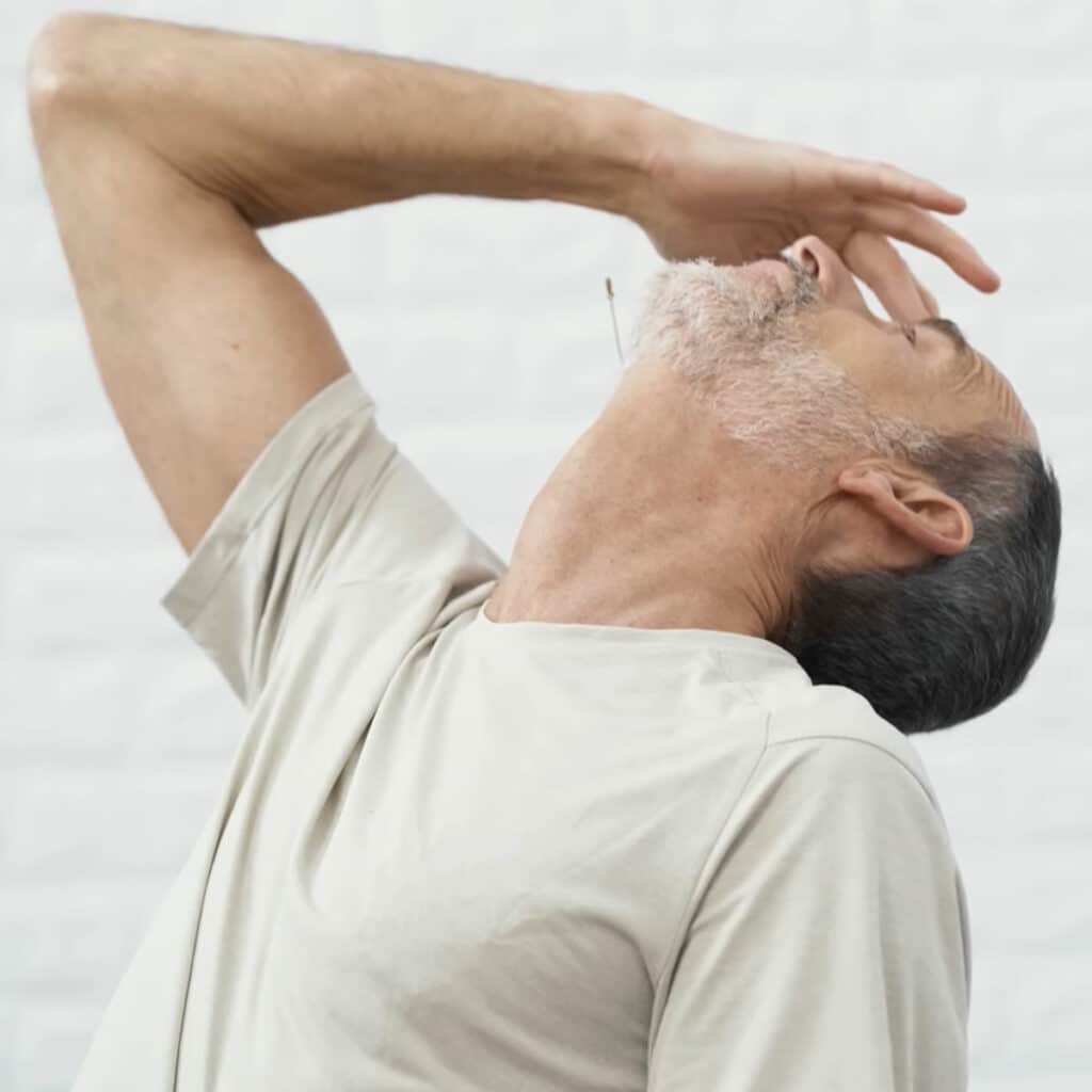 Roland Liebscher-Bracht legt seinen Kopf weit in den Nacken. Mit seiner rechten Hand tippt er seine Stirn an, um den Kopf noch weiter nach hinten zu bewegen.