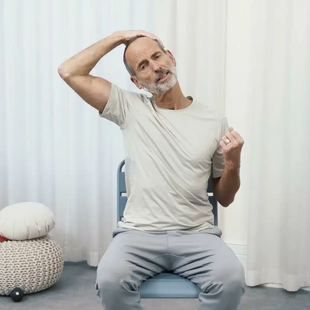 Roland Liebscher-Bracht sitzt aufrecht auf einem Stuhl. Seinen linken Arm hat er angewinkelt, die Hand zu einer Faust gebildet. Er zieht die Schulter runter, während er mit seiner rechten Hand an seinen Kopf greift und diesen zur rechten Seite zieht.