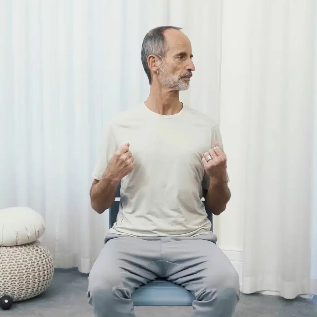 Roland Liebscher-Bracht sitzt aufrecht auf einem Stuhl. Seine Arme hat er angewinkelt. Die Hände sind leicht zu Fäusten geballt. Er zieht die Schultern runter. Seinen Kopf bewegt er zur linken Seite.