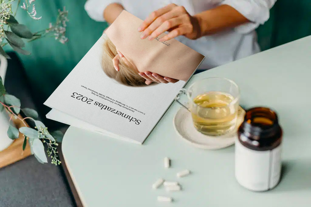 Eine Frau blättert im Schmerzatlas 2023 von Liebscher & Bracht. Vor ihr auf dem Tisch liegen Nahrungsergänzungsmittel ud eine Tasse Tee steht da.