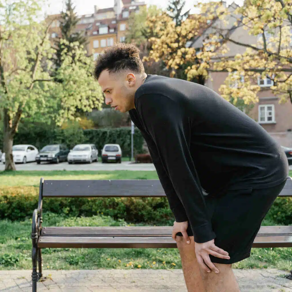 Eine Person steht vor einer Parkbank und hat die Hände auf die Knie abgestützt. Sie sieht erschöpft aus.