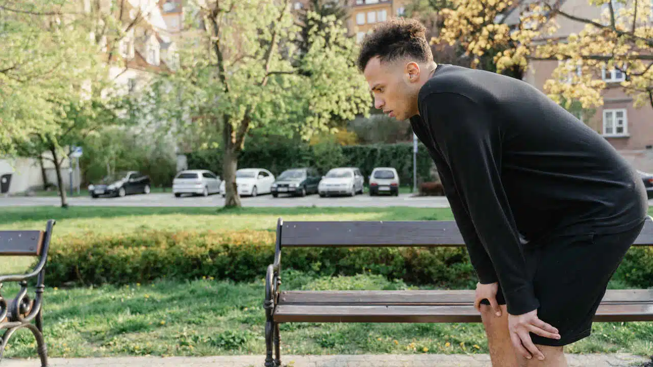 Eine Person steht vor einer Parkbank und hat die Hände auf die Knie abgestützt. Sie sieht erschöpft aus.