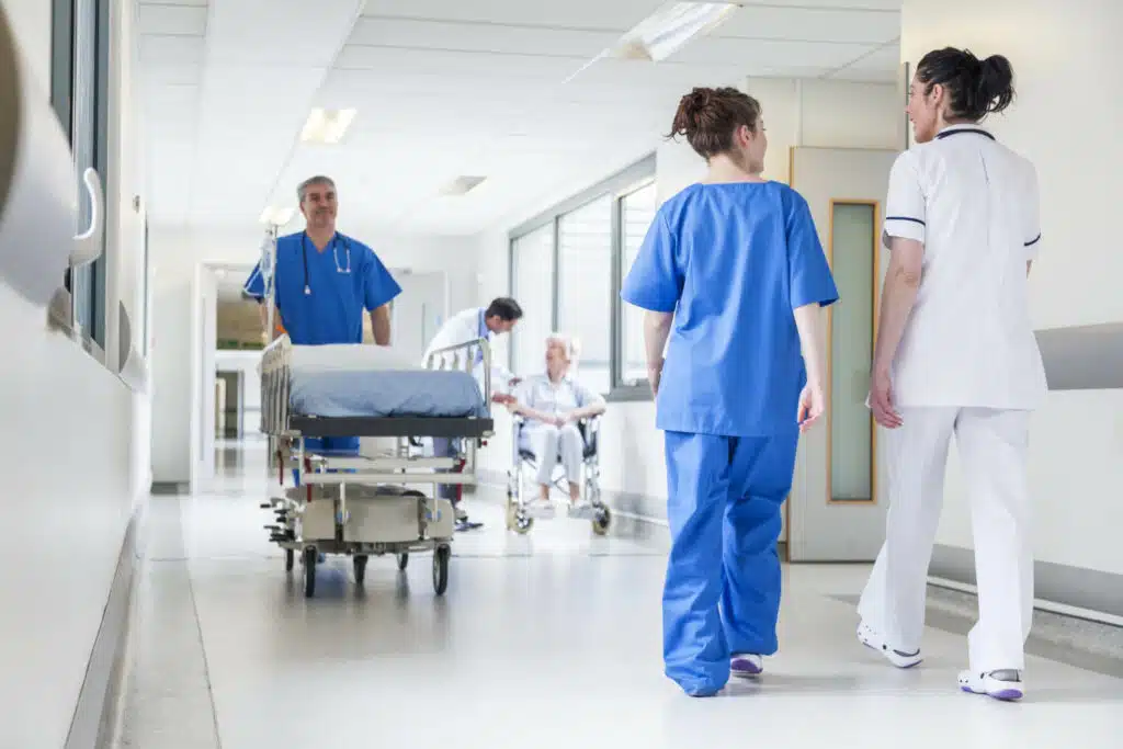 Pfleger schiebt Krankenhausbett durch Flur, Ärztinnen kommen ihm entgegen