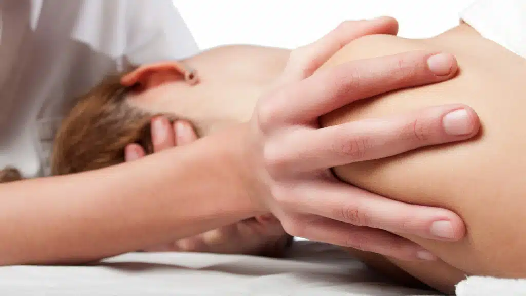 Ein Therapeut oder eine Therapeutin fixiert mit seiner Hand die Schulter seiner/ihrer Patientin, während er/sie gleichzeitig mit der anderen Hand den Kopf bewegt.