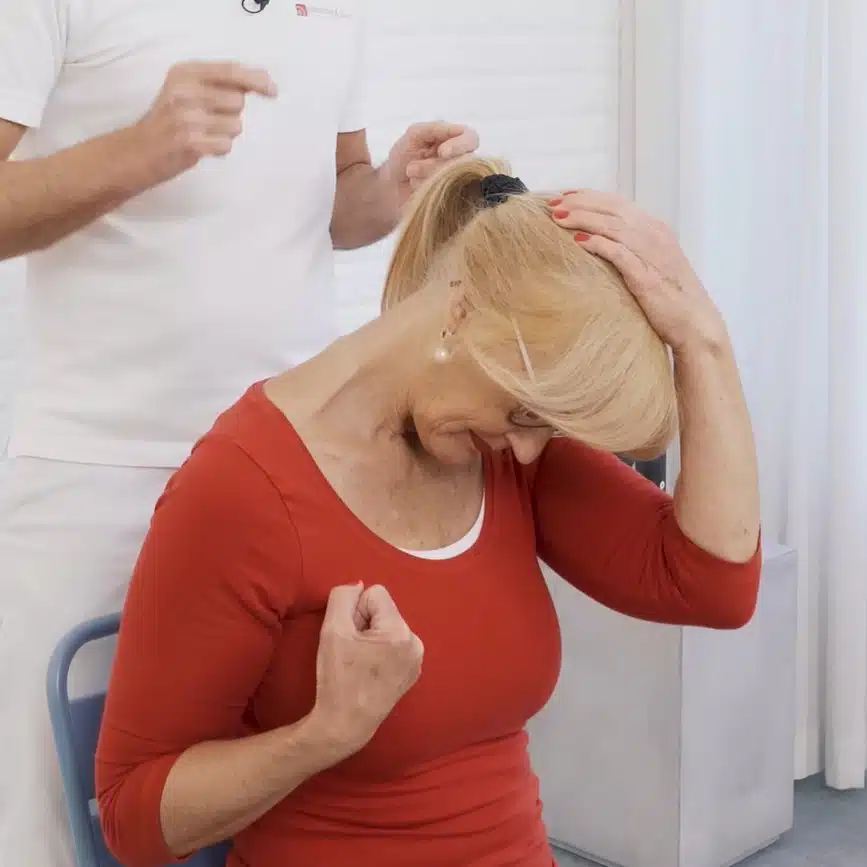 Eine Frau sitzt auf einem Stuhl. Ihre linke Hand liegt auf ihrem Kopf, ihren rechten Arm beugt sie an, um die Schulter nach unten zu ziehen. Den Kopf zieht sie nach links unten.