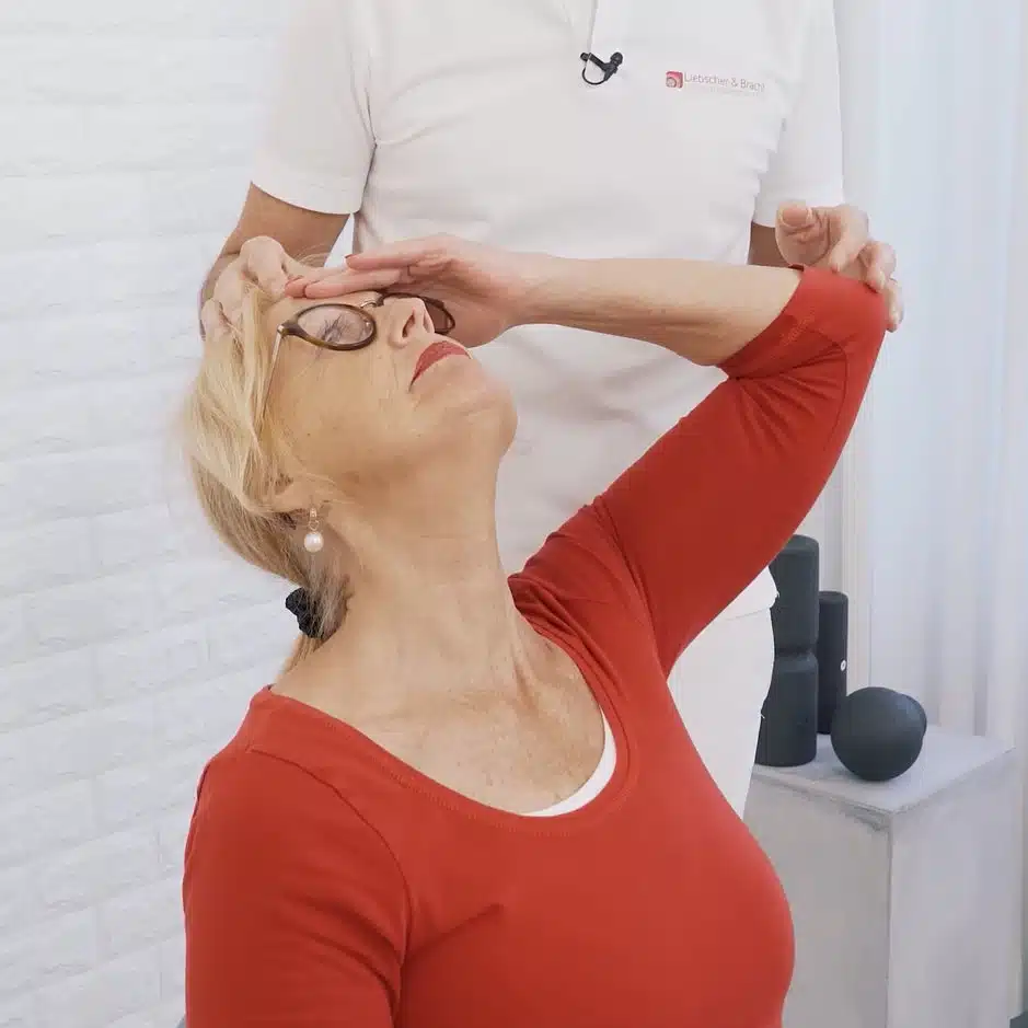 Eine Frau sitzt auf einem Stuhl, mit ihren linken Fingern tippt sie sich an die Stirn und legt ihren Kopf in den Nacken.