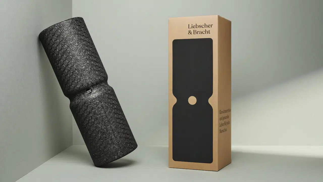Die Maxi-Faszienrolle von Liebscher & Bracht ist ganz schwarz und steht vor einem grau-grünem Hintergrund. Neben ihr steht der Karton, in dem sie geliefert wird. Auf dem Karton steht Liebscher & Bracht und eine schematische schwarze Faszienrolle ist abgebildet.