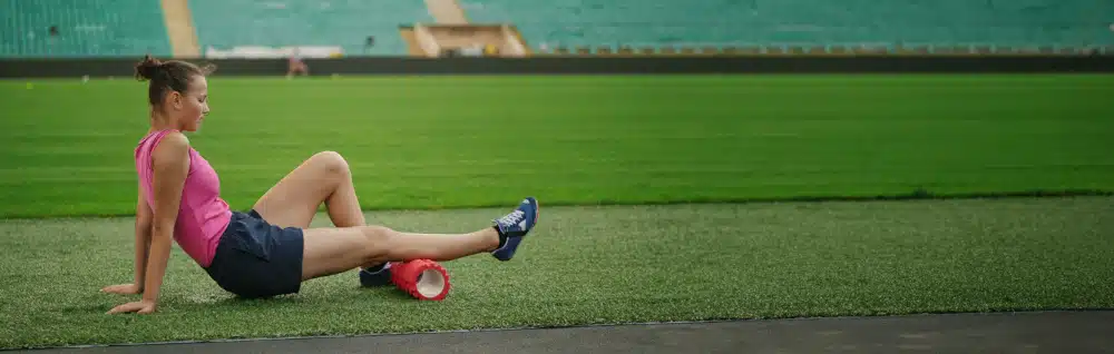 Frau in Sportkleidung sitzt auf Rasen in Stadion und rollt rechte Wade über eine Faszienrolle
