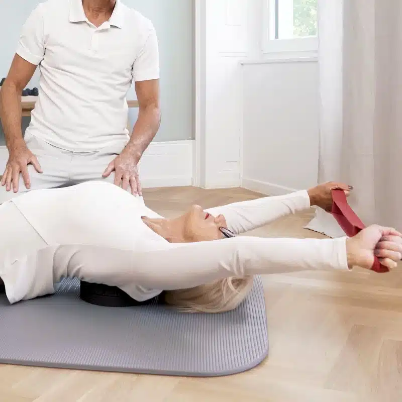 Eine Frau liegt am Boden auf einem Rückenretter von Liebscher & Bracht. Sie streckt ihre Arme gerade über den Kopf und hält mit beiden Händen eine rote Schlaufe.