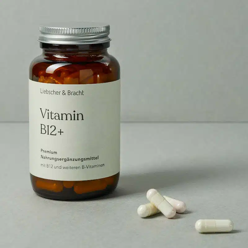 Eine kleine, dunkle Glasflasche steht vor einem dunkelgrünen Hintergrund. Sie ist mit Vitamin B12plus beschriftet. Daneben liegen vier Tabletten in heller Farbe.