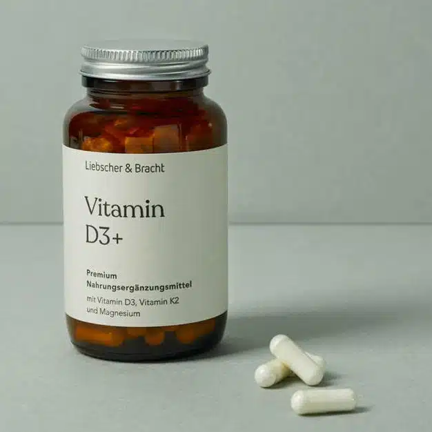 Eine kleine, braune Glasflasche steht vor einem olivgrünen Hintergrund. Sie ist mit D3plus beschriftet. Davor liegen drei weiße Pillen.