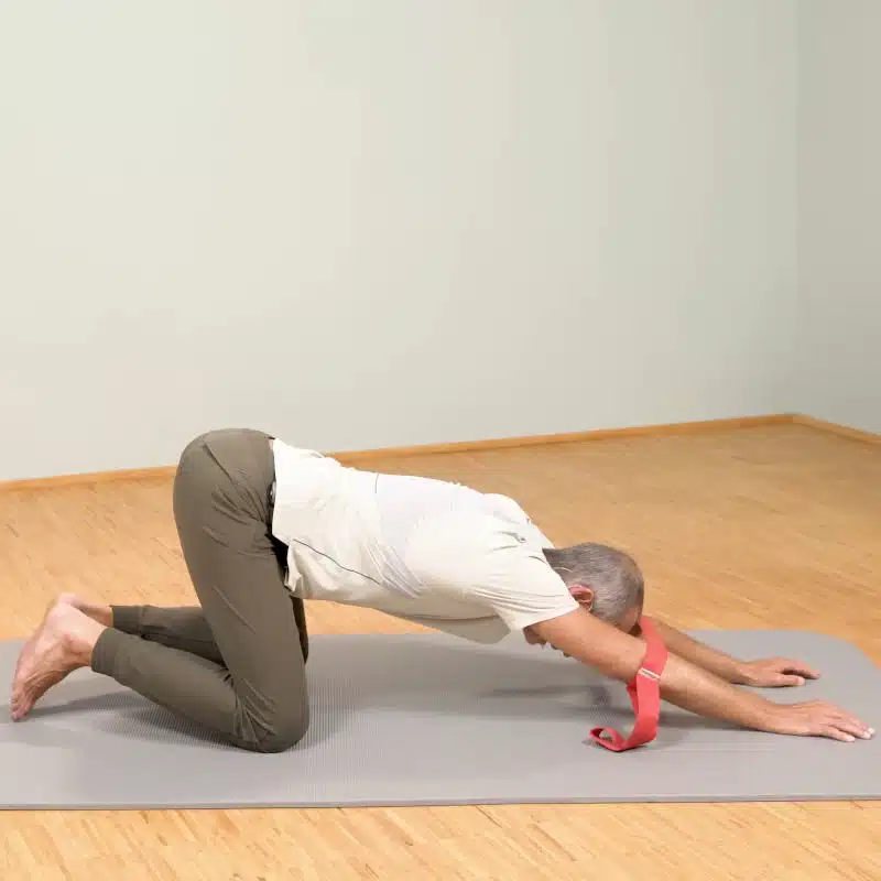 Roland Liebscher-Bracht kniet im Vierfüßlerstand am Boden. Er stützt sich mit den Händen weit vor seinem Körper am Boden ab und beugt sich mit der Brust hinunter. Um seine Ellenbogen hat er die Übungs-Schlaufe von Liebscher & Bracht gebunden.