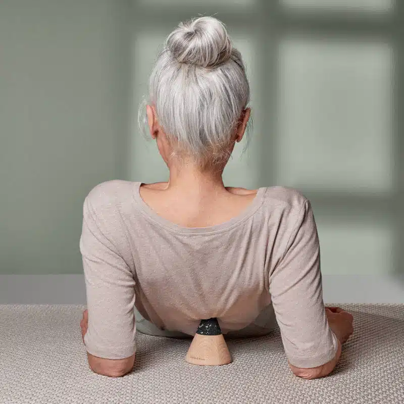 Eine Frau mit grauem Haar liegt am Boden auf einer beigen Matte. Sie stützt sich mit den Ellenbogen vom Boden ab. Ein kegelförmiger Schmerzfrei-Drücker ist unter ihrem Rücken platziert.