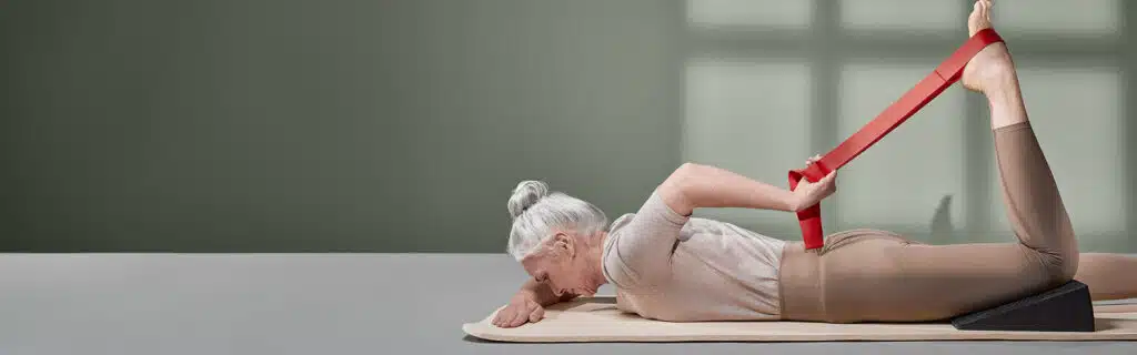 Eine Frau dehnt ihren Oberschenkel, indem sie auf dem Bauch liegend an der Übungsschlaufe zieht, die an ihrem Fuß befestigt ist. Unter dem Knie liegt zudem der Liebscher & Bracht Knieretter.