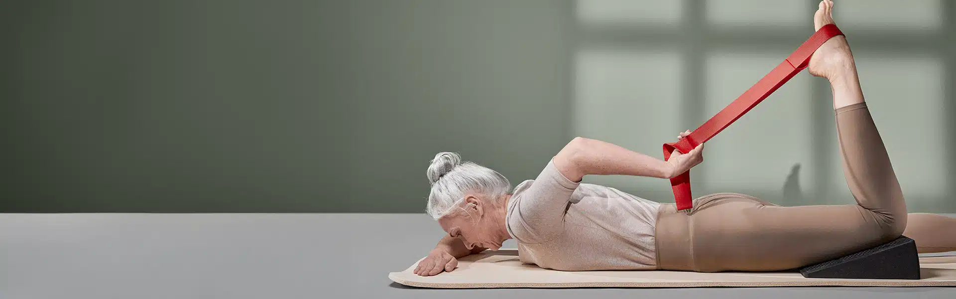 Eine Frau dehnt ihren Oberschenkel, indem sie auf dem Bauch liegend an der Übungsschlaufe zieht, die an ihrem Fuß befestigt ist. Unter dem Knie liegt zudem der Liebscher & Bracht Knieretter.