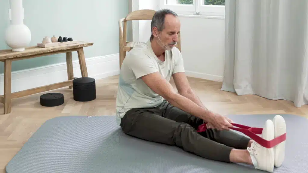 Roland Liebscher-Bracht sitzt auf dem Boden und hat die Beine nach vorne gestreckt. Die Übungsschlaufe liegt um seine Füße und er zieht den Rumpf nach vorne Richtung Füße.