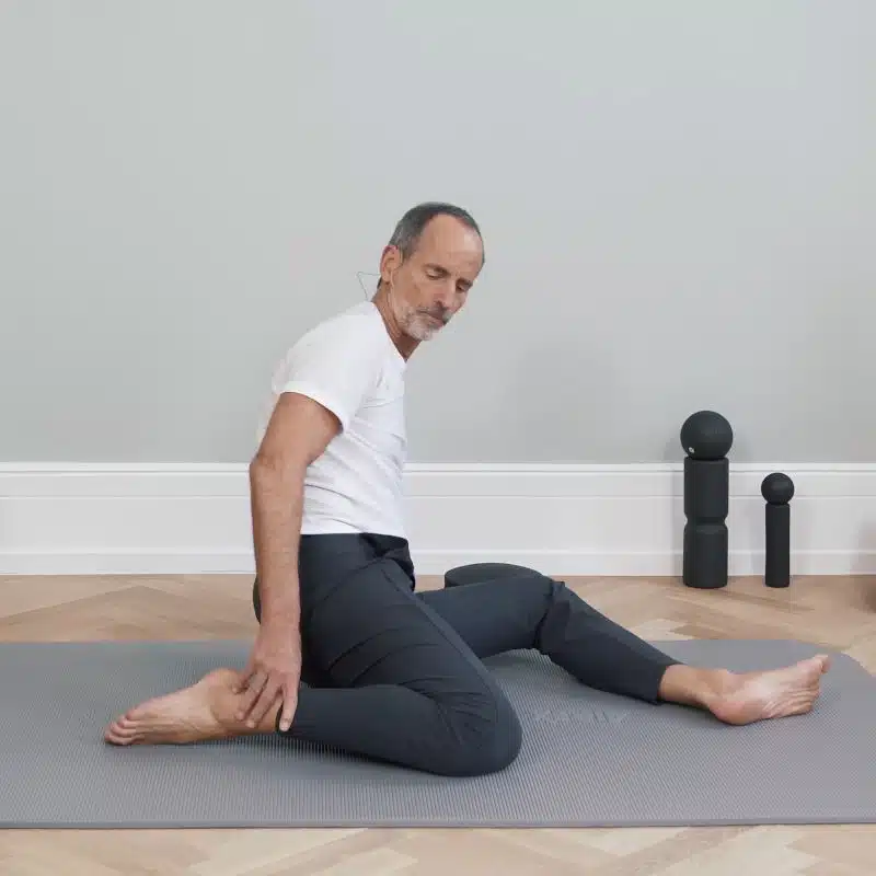 Roland Liebscher-Bracht sitzt am Boden auf einer grauen Übungsmatte. Sein linkes Bein liegt vor ihm, während er den rechten Fuß mit einer Hand zu seinem Gesäß zieht.