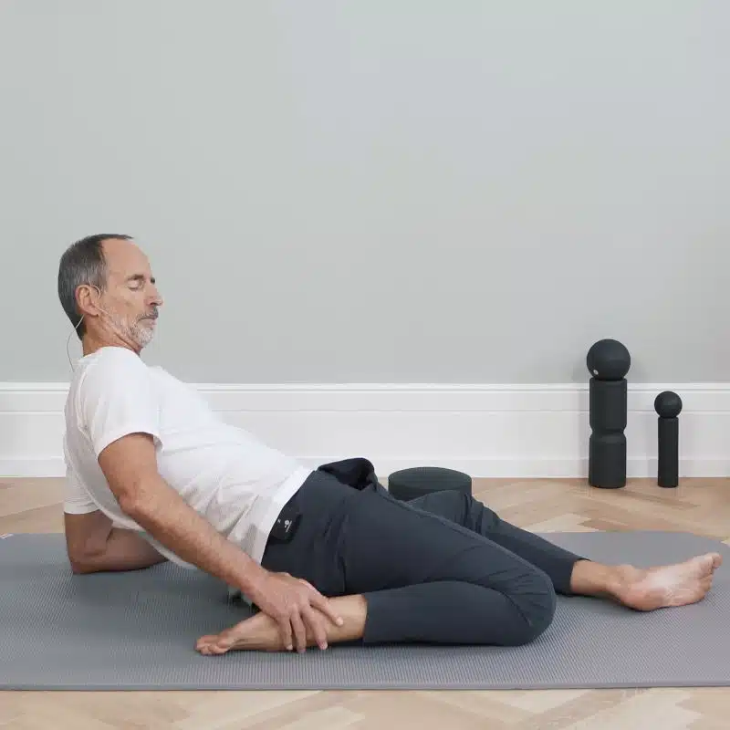 Roland Liebscher-Bracht macht am Boden eine Übung. Dazu hat er sein linkes Bein nach vorne ausgestreckt, während das rechte Bein angewinkelt ist und neben seinem Gesäß liegt. Mit dem Oberkörper hat sich Roland nach hinten gebeugt und stützt sich auf seinem Unterarm ab.