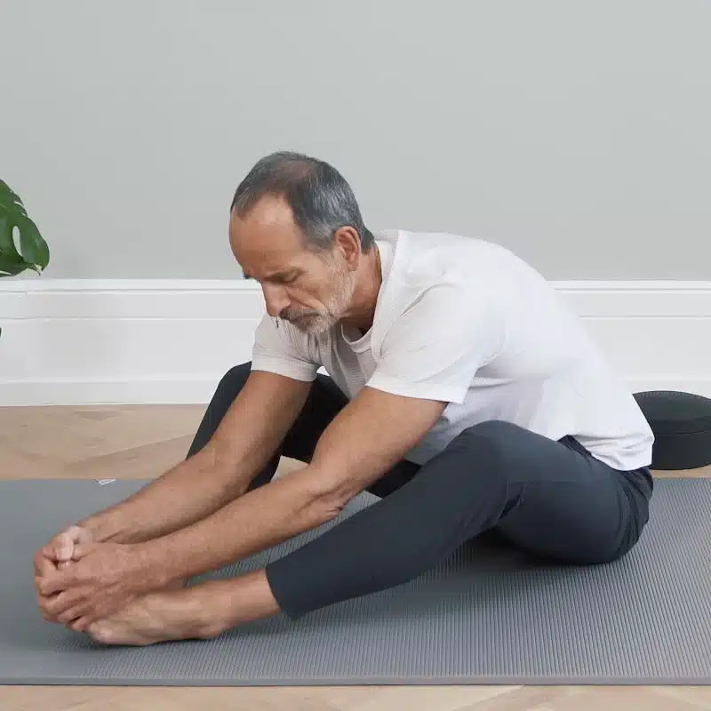 Roland Liebscher-Bracht sitzt am Boden und beugt sich mit dem Oberkörper nach vorne über seine fast ausgestreckten Beine. Er hält sich mit beiden Händen an seinen Füßen fest.
