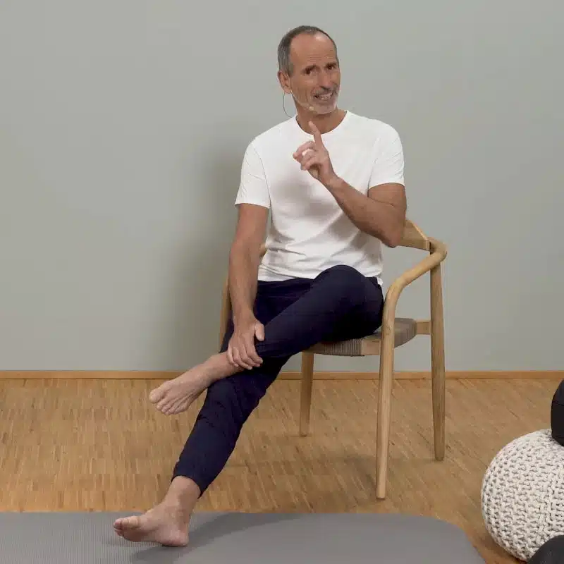 Roland Liebscher-Bracht sitzt auf einem Holzstuhl. Er hat das linke Bein leicht über sein rechtes Knie gelegt und schaut in die Kamera.