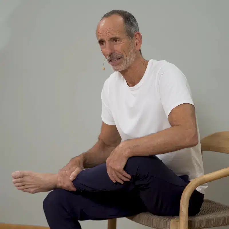 Roland Liebscher-Bracht sitzt auf einem Holzstuhl. Er hat den linken Fuß auf dem rechten Oberschenkel abgelegt und beugt seinen Oberkörper nach vorne.