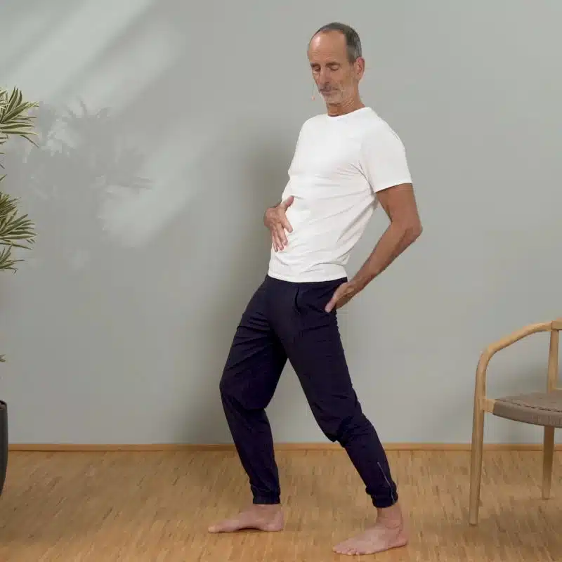 Roland Liebscher-Bracht schiebt für einen Übung seine Leiste in Schrittstellung nach vorne. Er hat seine Hände auf Bauch und Gesäß platziert.