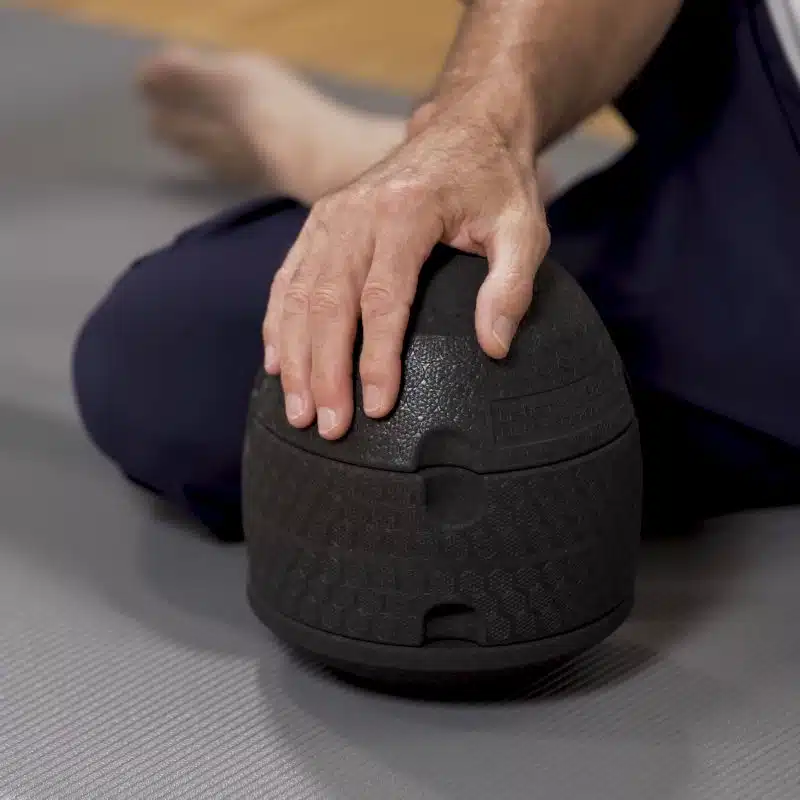 Der ISG-Ischias-Retter von Liebscher & Bracht steht auf einer grauen Yogamatte, während eine Person ihn mit einer Hand hält.