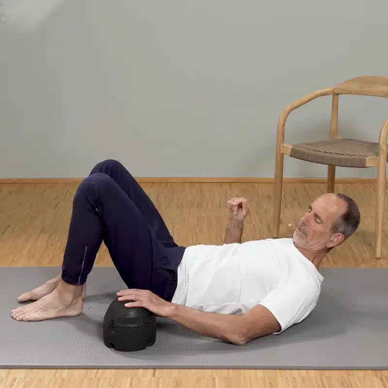 Roland Liebscher-Bracht liegt mit dem Rücken auf einer grauen Trainigsmatte. Neben ihm steht der ISG-Ischias-Retter bereit.