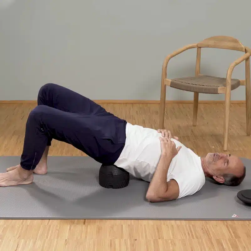 Roland-Liebscher-Bracht liegt mit seinem Iliosakralgelenk auf dem ISG-Ischias-Retter. Seine Beine sind dabei angewinkelt und die Füße aufgestellt.