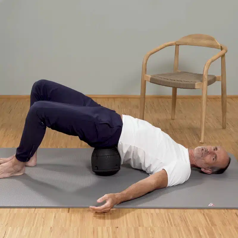 Roland Liebscher-Bracht massiert sein Kreuzbein-Darmbein-Gelenk, indem er auf dem ISG-Ischias-Retter liegt und kreisende Bewegungen macht. Dabei sind seine Füße aufgestellt, die Beine angewinkelt und die Arme seitlich am Boden ausgestreckt.