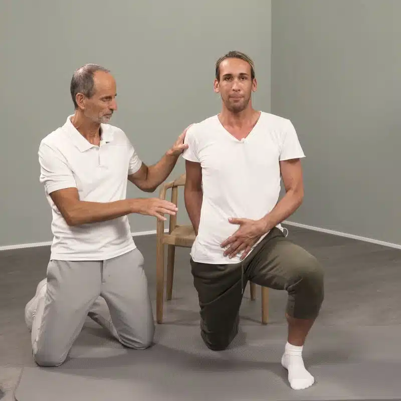 Roland Liebscher-Bracht kniet neben einem ebenfalls knienden Mann, der sein linkes Bein für eine Übung nach vorne aufgestellt hat.