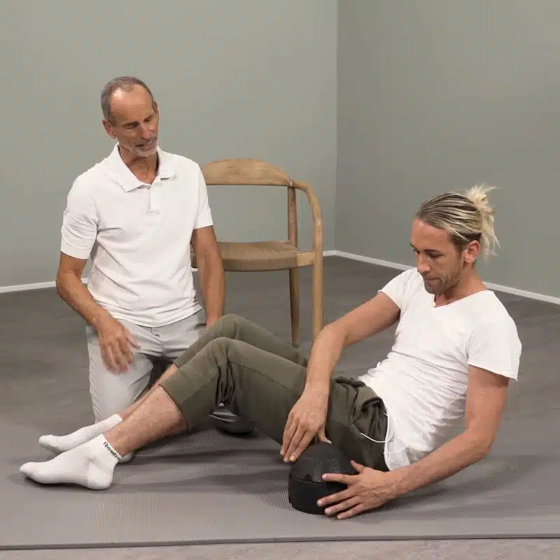 Roland Liebscher-Bracht schaut zu, wie ein Mann versuchst, sich am Boden auf den ISG-Ischias-Retter zu legen.