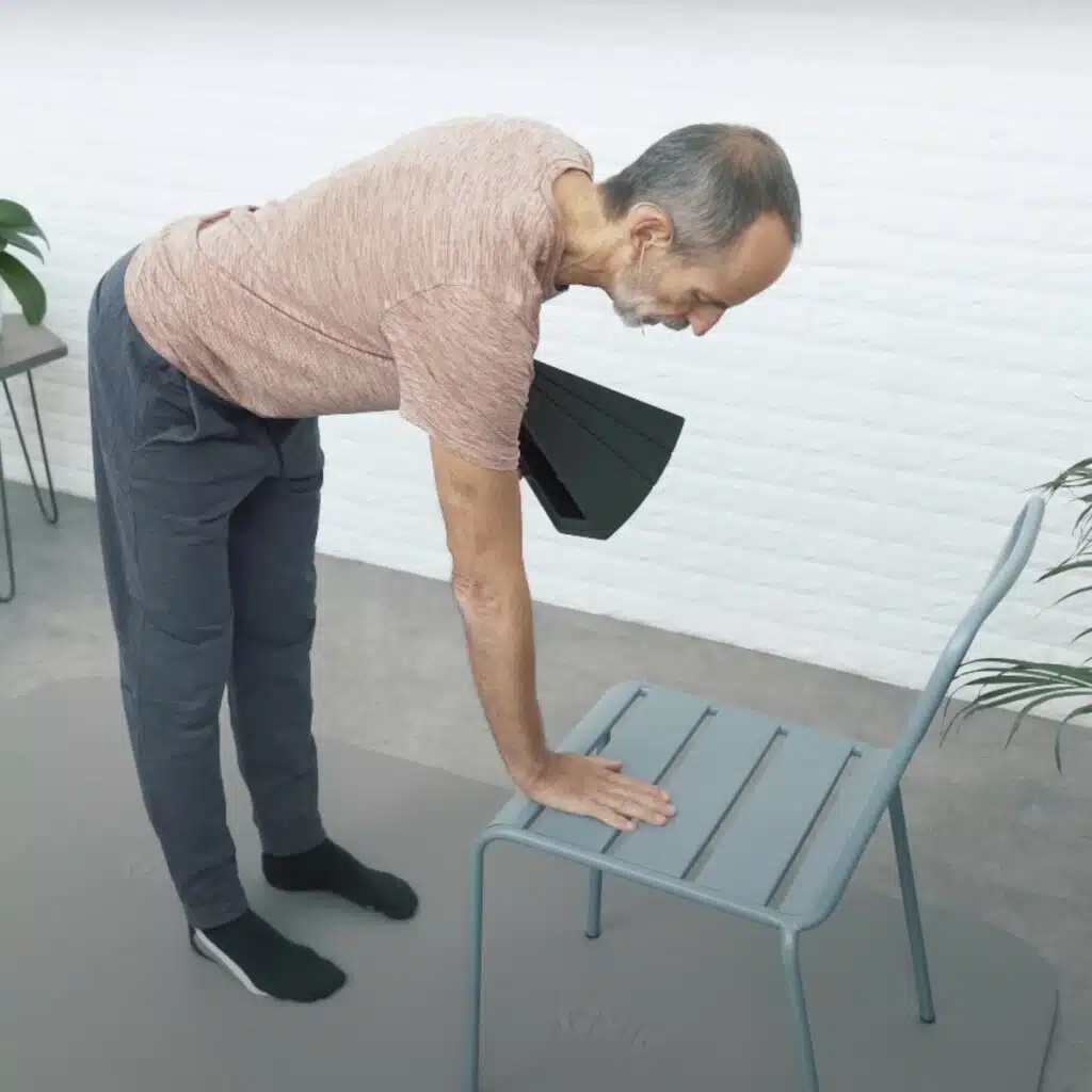Eine Person beugt sich über einen Stuhl, um sich darauf abzustützen. Der Oberkörper ist parallel zum Boden, die Beine sind gestreckt. In der Hand hält sie den Knieretter, um die Übung zu intensivieren.