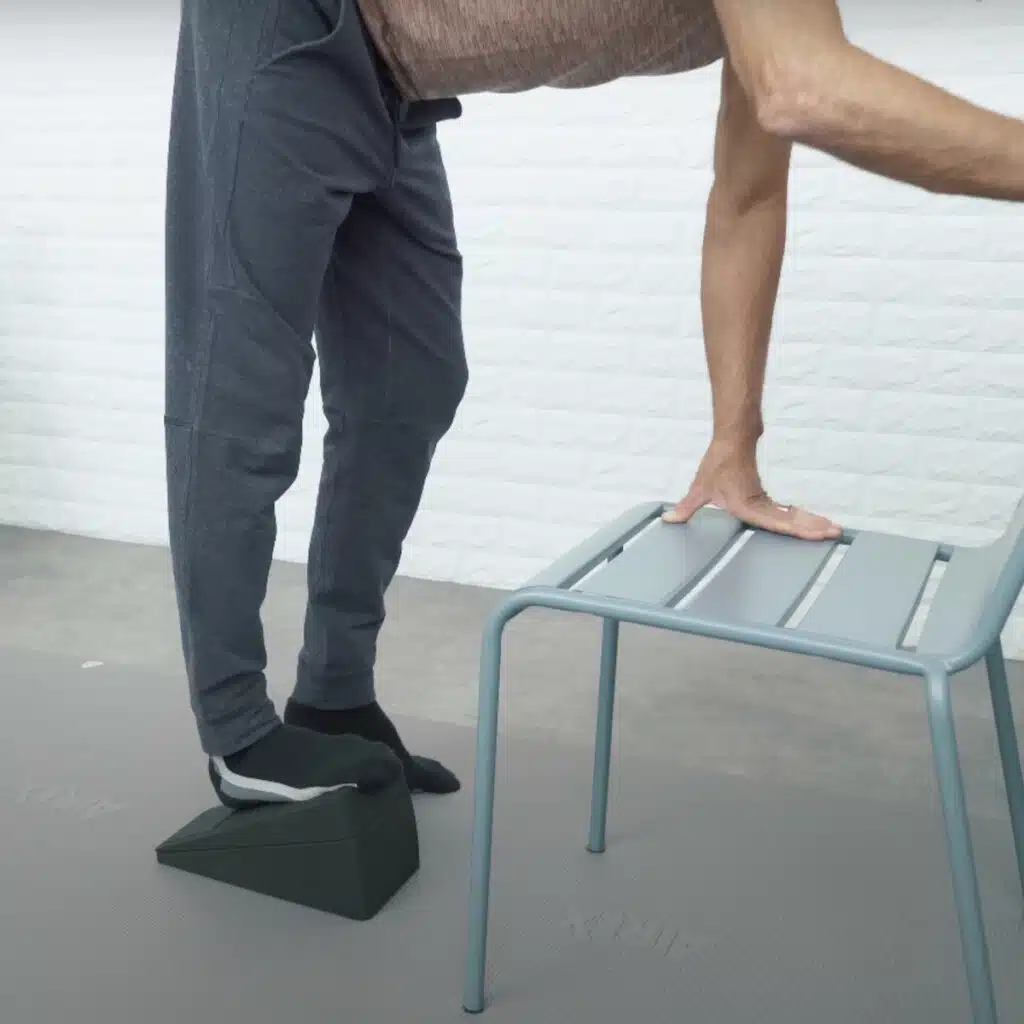 Eine Person steht vor einem Stuhl und stützt sich mit den Händen am Stuhl ab. Sie ist mit dem Oberkörper nach vorne geneigt und hat den rechten Fuß auf dem Knieretter abgestellt.