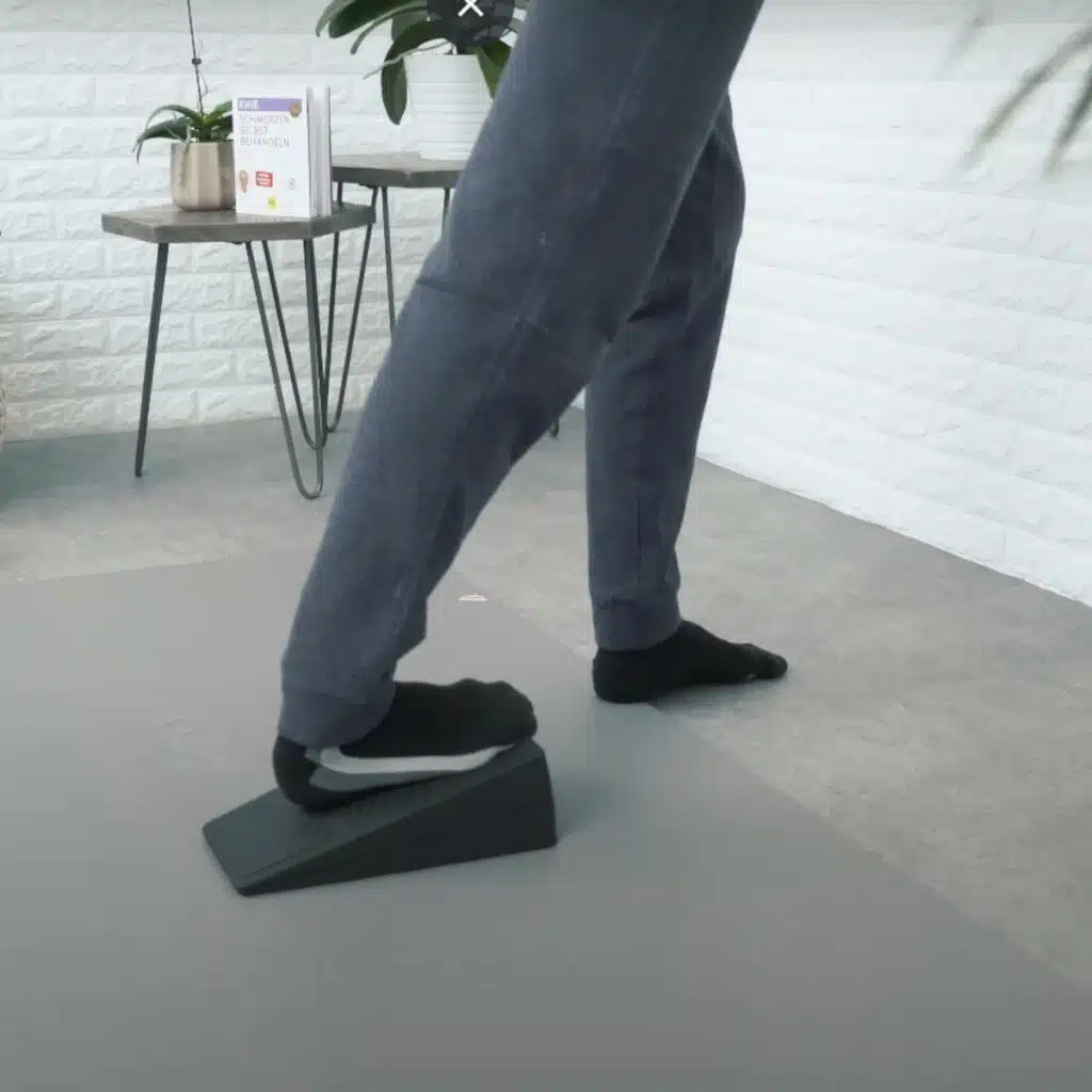 Eine Person steht vor einer Wand und steht mit dem rechten Fuß auf dem Knieretter von Liebscher & Bracht. So wird die Wade gedehnt gegen eine Bakerzyste.