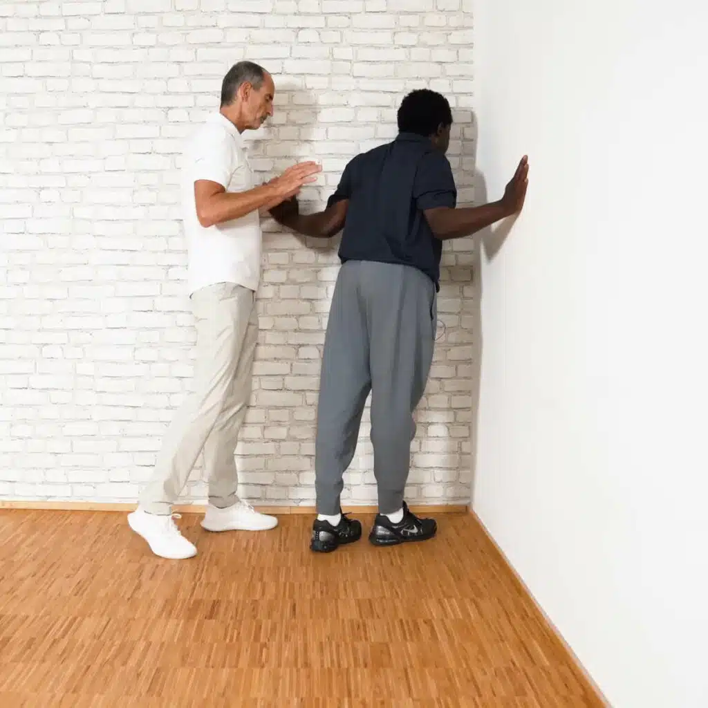 Eine Person steht mit angewinkelten Armen in einer Raumecke. Die Handflächen berühren die Wände rechts und links, die Ellenbogen sind eng am Körper angewinkelt. Die Person lehnt ihren Oberkörper in die Ecke.