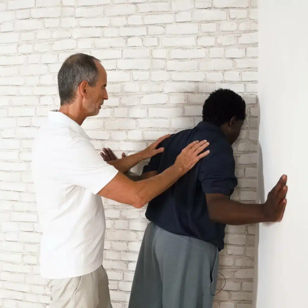 Eine Person lehnt den Oberkörper in eine Raumecke, dabei sind die Arme seitlich angewinkelt, sodass die Ellbogen eng am Körper sind und die Unterarme zu den Seiten weggestreckt. Beide Hände berühren die Wände. Roland Liebscher-Bracht hat seine Hände auf den Rücken der Person gelegt und signalisiert das Nach-vorne-Gehen mit dem Oberkörper.