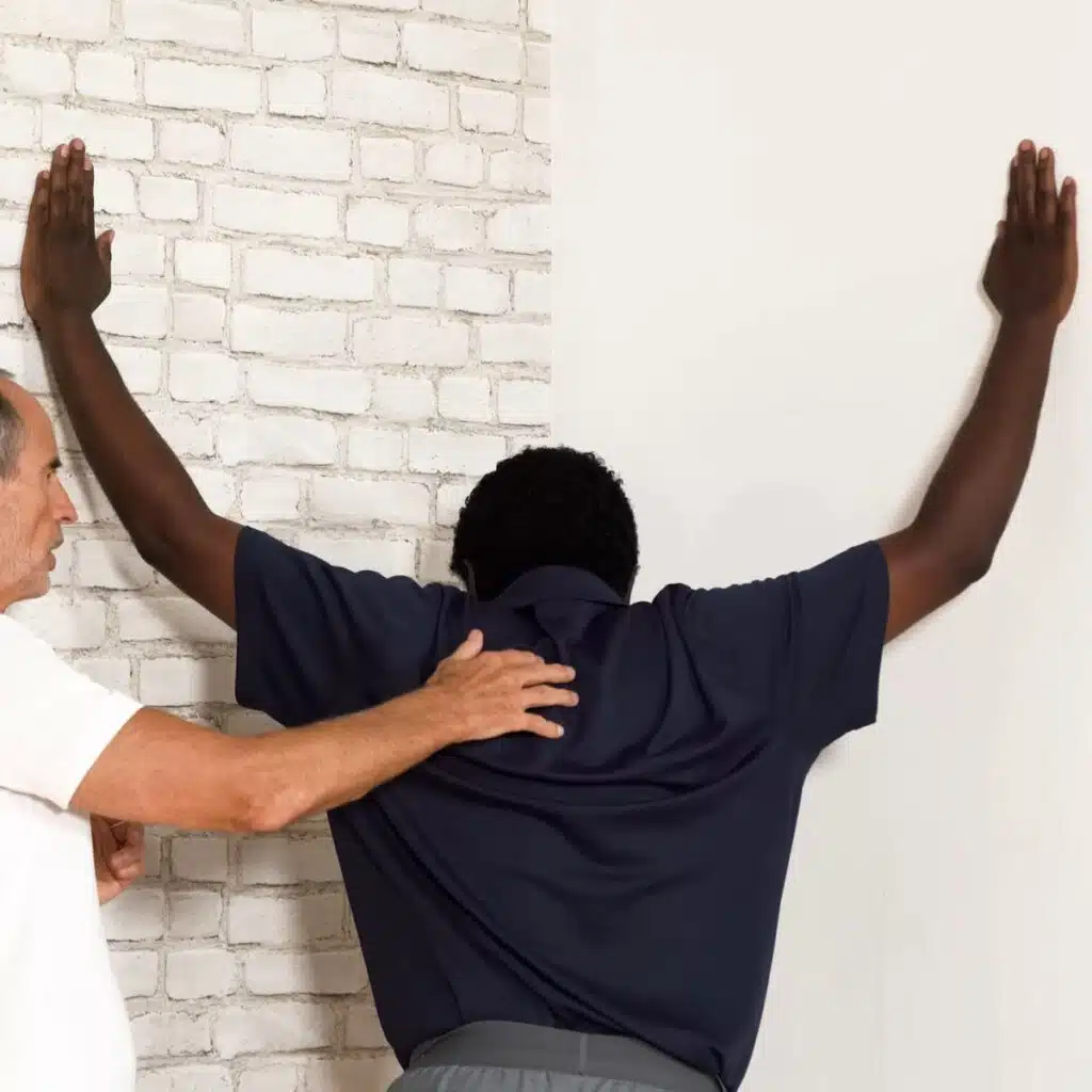 Eine Person steht in einer Raumecke und hat beide Arme zu den Seiten angehoben und angewinkelt. Die Handflächen liegen an den Wänden rechts und links. Sie lehnt den Oberkörper in Richtung Ecke nach vorne.