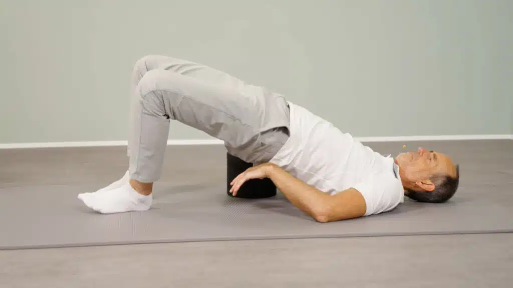 Roland Liebscher-Bracht liegt mit dem Rücken auf einer Gymnastikmatte. Er hat die Füße aufgestellt und sein Becken in die Luft angehoben. Mit den Händen greift er den Rückenretter und platziert ihn unter seinem Kreuzbein.