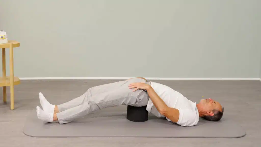 Roland Liebscher-Bracht liegt auf einer Gymnastikmatte auf dem Rücken. Unter seinem Kreuzbein befindet sich der Rückenretter, sodass sein Becken nach oben angehoben wird. Seine Schultern, sein Kopf und seine Füße berühren den Boden.