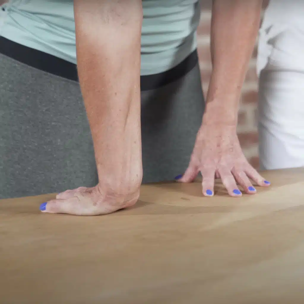 Eine Person hat die Hände auf einen Tisch abgestützt. Sie dreht die rechte Hand nach hinten, sodass die Finger zu ihr schauen.