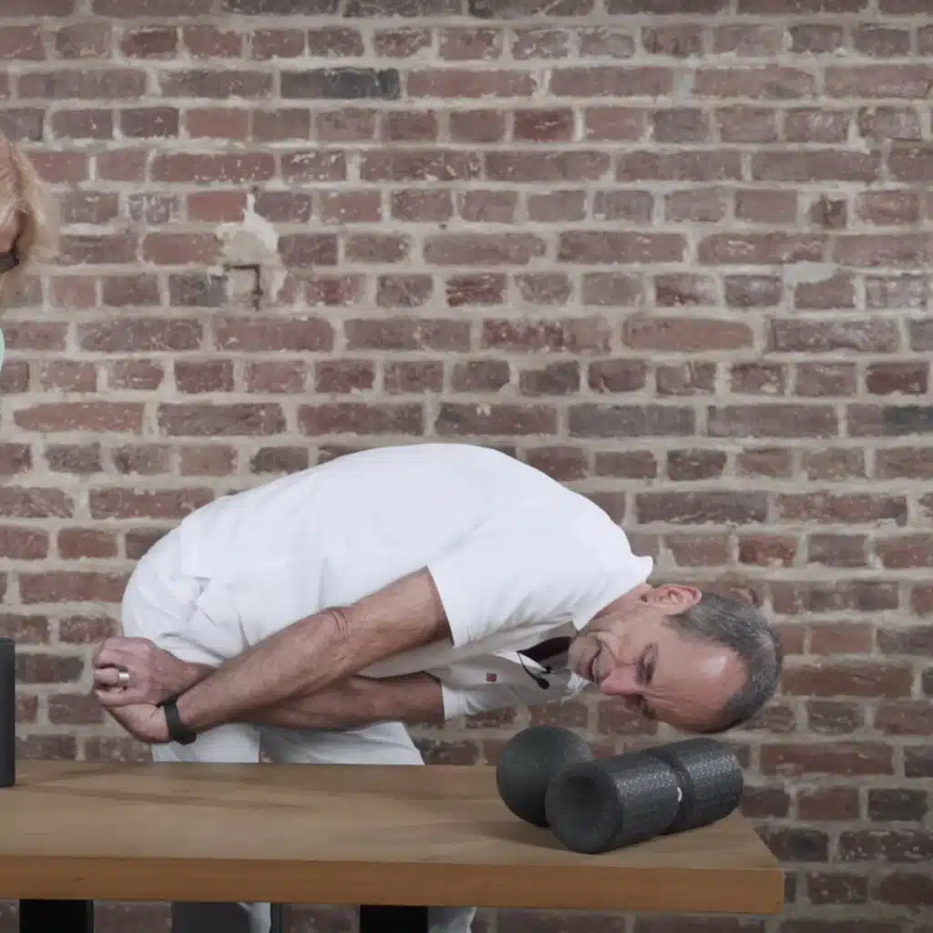 Eine Person neigt sich über einen Tisch, um eine Dehnung auszuführen. Sie hat mit der linken Hand die rechte Hand gegriffen. Die rechte Hand ist zum Unterarm eingerollt, der rechte Arm ist in einer Innenrotation.